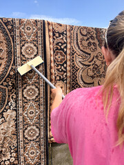 A young woman in a pink T-shirt with tied-back hair is cleaning a hanging carpet outdoors using a brush.