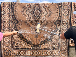 A traditional carpet being washed outdoors with a hose and scrub brush. A woman's hand and a man's hand are seen from opposite sides, actively cleaning the rug with water.