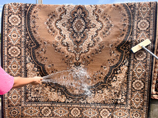 A traditional carpet being washed outdoors with a hose and scrub brush. A woman's hand and a man's hand are seen from opposite sides, actively cleaning the rug with water.