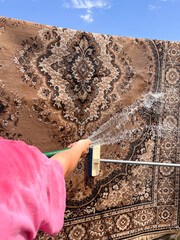 A vertical shot of a woman’s hand in a pink t‑shirt holding a garden hose spraying water onto a carpet being washed outdoors.