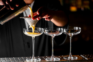 Bartender pouring cocktail through strainer into coupe glass at bar. Professional mixology, precise technique, dramatic lighting, upscale nightlife concept.
