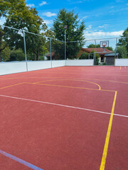 Fototapeta premium Basketball court with vibrant red surface showcases clear blue sky and green trees in a quiet park setting during midday