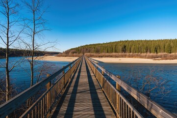 Naklejka premium Pedestrian wooden footbridge crossing a river surrounded by forest under a clear blue sky in late winter.
