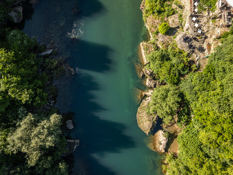 Top down aerial photo of the turquoise Neretva River with rocks and trees on both banks, the river flowing vertically, and a glimpse of Restaurant Babilon near the Old Bridge