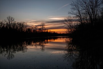 Obraz premium Evening scene of a broad river's opposite bank featuring leafless tree shadows mirrored on the water at sunset.