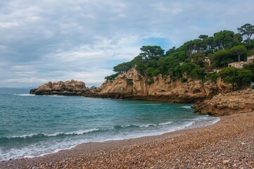 Morning view of a sandy and pebble shore nestled in a rocky cove beneath umbrella pines by the sea.