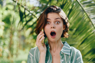 Young woman in a striped shirt talks on a mobile phone outdoors, eyes wide with surprise as sunlight filters through palm leaves.