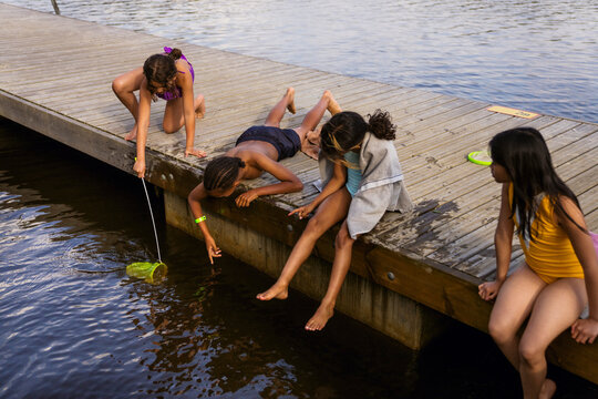 Girl catching fish with net in lake near boy lying on pier at summer camp