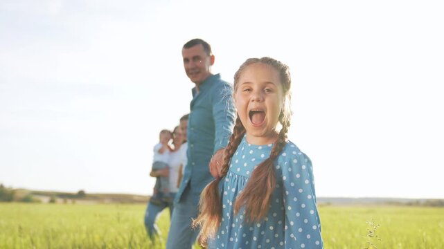 Family walking hand in hand across sunlit meadow with father and mother leading daughter and son while parents smile and child laughs in polka dot dress on green grass under warm sun full of joy