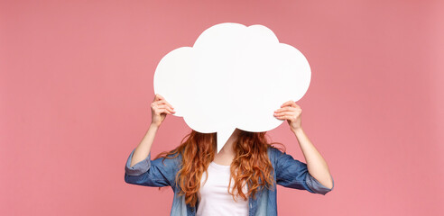 Unrecognizable redhead female holding an empty speech bubble in front of her head, pink studio background, panorama with empty space