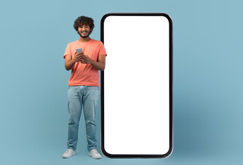 Online offer concept. Happy curly young indian man using cell phone on blue studio background, standing by big mobile phone with white empty screen, full length shot, panorama with copy space, mockup