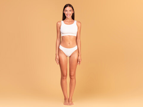 Full length frontal view of slim woman with perfect body shape and flat tummy posing on beige studio background, lady wearing white lingerie, copy space