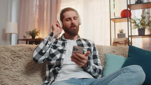 Young man chills at home with wireless headphones listening music dancing, bobbing head to a catchy beat. Caucasian guy on sofa lifts thumb up, indicating approval of an awesome song tune in rotation