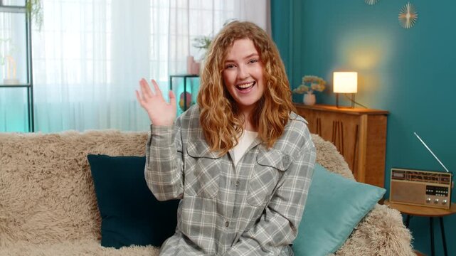 Young woman at home smiling warmly and waving hand to say hello with friendly and cheerful energy. Caucasian redhead girl on sofa expressing joy, welcoming gesture and positive emotional connection