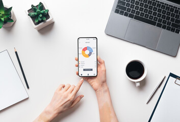 Business lady hands holding mobile phone with trading app over workdesk, top view, creative image. Unrecognizable female manager using financial application on smartphone while working in office