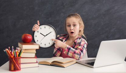 Portrait of surprised smart girl sitting with stack of books and laptop, holding big clock and pointing on it. Education, development, time management, deadline, time to study, school concept