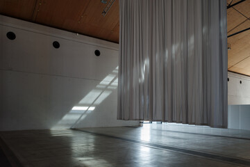 Minimalist architectural interior of a large, empty exhibition hall or gallery space in Berlin. A massive grey curtain hangs from the high wooden ceiling, and bright sunlight casts strong diagonal