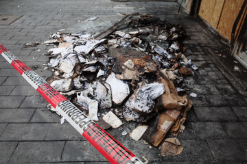 The destructive aftermath of a fire on a city sidewalk in Berlin, Germany. A pile of burnt paper and debris lies on the pavement, cordoned off by red and white 'Polizeiabsperrung' (Police Cordon) tape