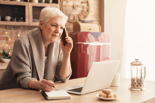 Senior Businesswoman Using Laptop, Talking On Cellphone And Taking Notes While Working From Home Sitting In Kitchen