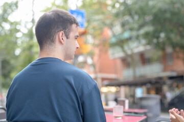 Fototapeta premium A man in a blue shirt is sitting at a table with a cup in front of him. The scene is set in a city with a blue sign in the background. The man is looking off into the distance