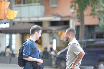 Two men are talking on a street corner. One of them is wearing a backpack. The other man is wearing a gray shirt