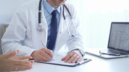 A doctor and a patient. The physician, wearing a white medical coat over a blue shirt and tie, is filling out a medical record form during a consultation in the clinic. Medical service