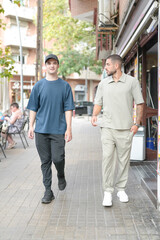 Two men walking down a street, one wearing a blue shirt and the other wearing a tan shirt