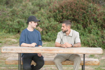 Two men are sitting on a wooden bench in a park. One of them is wearing a black hat