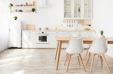 Scandinavian kitchen interior with white furniture and shelves, wooden dining table and small refrigerator near window, empty space. Dining room with sunbeams on the floor