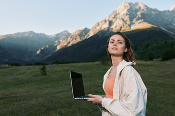 A confident woman stands outdoors with a laptop in hand, set against a scenic mountain landscape. She gazes ahead with calm determination, embodying remote work in nature.