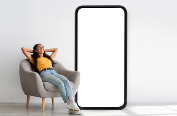 Happy young woman relaxing in armchair nar big smartphone with blank screen, offering space for app or website mockup near white studio wall. Cellphone display template, mobile application