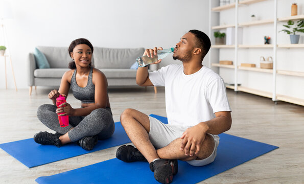 Athletic black couple in sportswear sitting on yoga mats, drinking water from bottles after domestic training, indoors. Cool guy and his girlfriend taking break after workout, staying hydrated