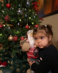 Cute little girl peeking from behind a decorated Christmas tree at home. Festive holiday atmosphere with lights, ornaments, and joyful winter mood. Perfect image for Christmas, New Year