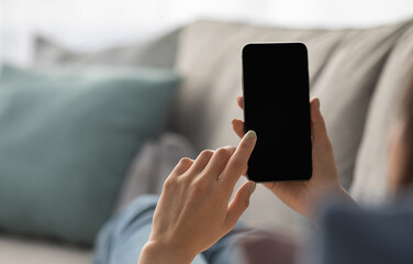 Woman relaxing at home on couch and surfing on internet in free time. Young lady uses smartphone with empty screen, typing message, watch video in living room interior, view over shoulder, cropped