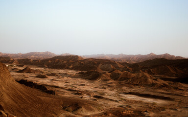 Fototapeta premium Vast arid desert landscape with undulating brown hills and distant mountain range under a clear, pale sky, evoking a sense of isolation and natural beauty.
