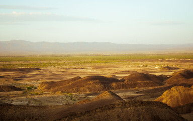 Expansive arid landscape with rolling brown hills and distant green valley under a soft hazy sky, evoking a sense of vastness and natural beauty