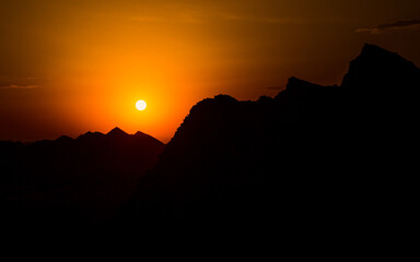 Stunning sunset paints the sky in vibrant orange hues, silhouetting dramatic mountain peaks against the warm, fading light, a moment of serene natural beauty.