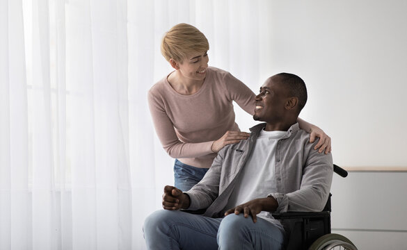 Medical equipment in hospital for assistance handicapped people and help. Smiling woman nurse care about smiling adult african american disabled guy in wheelchair, in living room or clinic interior - Powered by Adobe