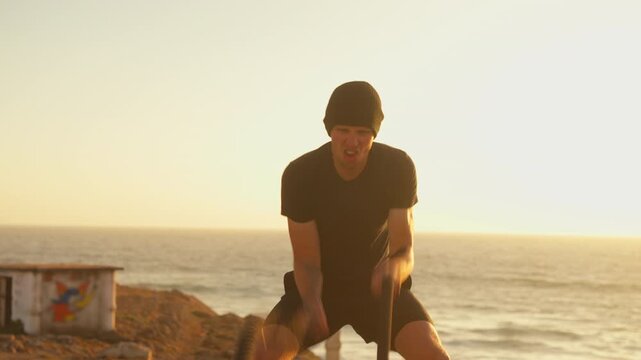 Athletic man training with battle ropes by sea during sunset. Sport, fitness, and healthy lifestyle concept. Caucasian sportsman with muscular body focused on functional training and endurance 
