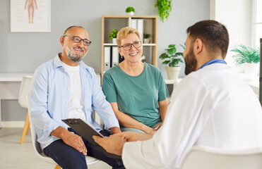 A senior couple smiles while talking with their family doctor during medical exam and consultation in bright clinic office, discussing treatment. They are attentive, comfortable and reassured.