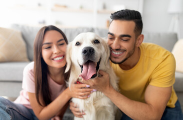 Portrait of happy multiracial couple hugging adorable dog, cuddling with their pet at home. Arab man and his Caucasian girlfriend embracing golden retriever, having fun times indoors, selective focus