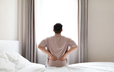 Tired man suffering from back pain after sleeping in bedroom, waking up in morning, sitting on bed...