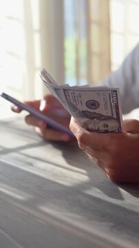 Close-up of man's hand holding smartphone and hundred dollar bills while taking notes, money financial management concept. Soft focus, Selective focus
