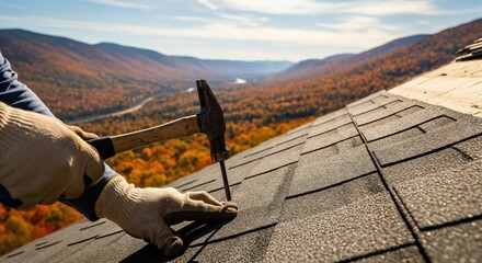 Close-up of a roofer's gloved hands hammering a nail into asphalt shingles on a house roof with a beautiful autumn mountain landscape in the background.