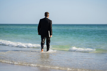 Businessman tourist in casual suit barefoot walking on tropical beach with laptop. Handsome business man on summer vacation. Businessman running on sea holding laptop. Businessman jumping in sea.