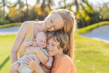 Fototapeta premium Mother, older son, and newborn baby on a meadow in the park. Family spending quality time together in nature. Parenthood, love, and family bond concept