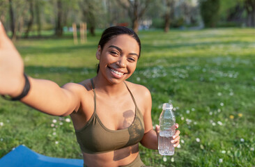 Cheery young black female vlogger taking selfie or speaking to followers remotely on mobile phone, doing yoga or meditation at city park. Sports blogger, online coach and distance training
