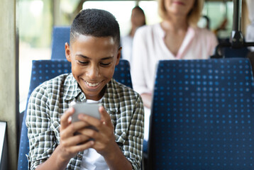 Happy black kid using cell playing mobile games online on smartphone connected to public wifi sitting on seat in city bus. Smiling teen passenger looking at device gadget screen, texting sms