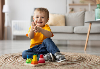 Adorable toddler boy playing with educational wooden toy at home, clapping hands and smiling at camera, sitting on carpet in living room. Cute child using colorful sorting stacking blocks puzzles