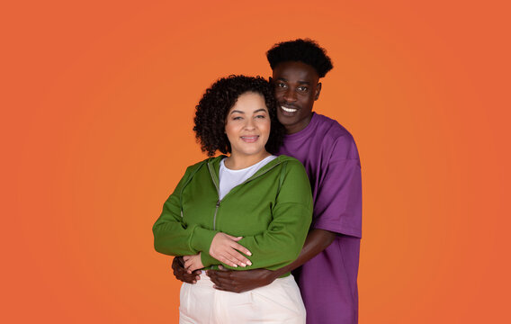 Studio portrait of cheerful beautiful young loving mixed race couple in casual hugging and smiling at camera on red background, african american guy embracing hispanic chubby woman, copy space